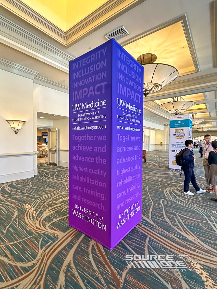 Tall purple branded tower display with sponsor messaging and conference information positioned in a convention center corridor.