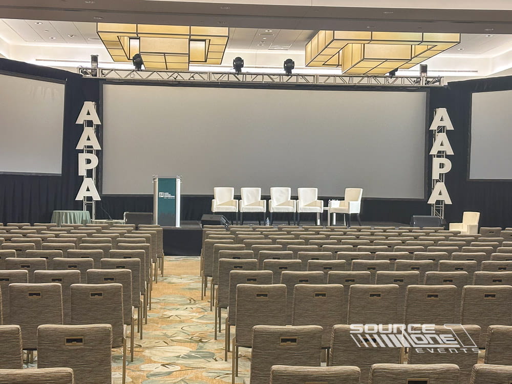 Professional conference stage with large projection screen, white presenter seating, and clean contemporary design.