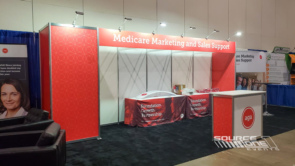 Healthcare trade show booth with bold red graphics, illuminated branding, reception counter, and exhibitor meeting space.