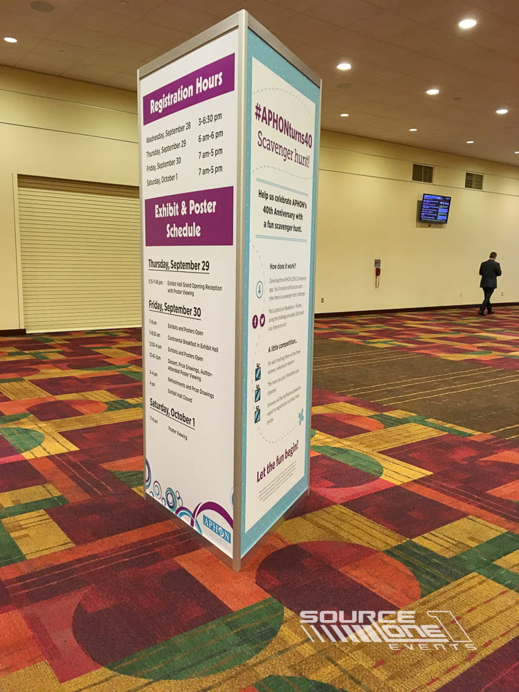 White freestanding tower display with colorful conference schedule graphics positioned in convention center corridors.