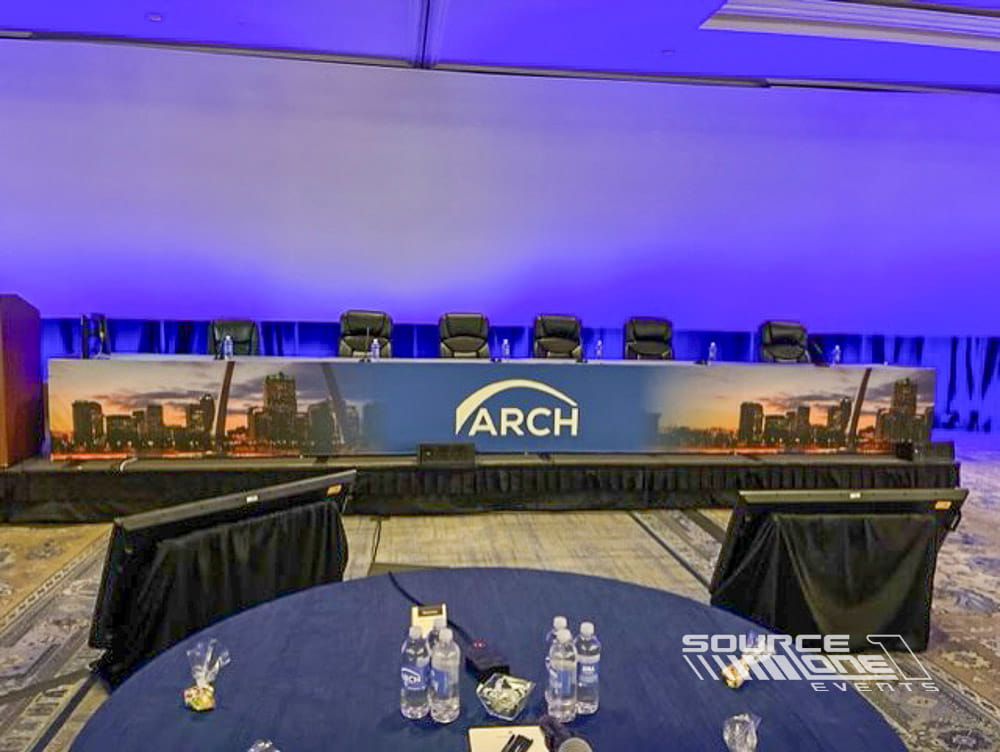 Contemporary conference stage with blue ambient lighting, curved presentation screens, and panel discussion seating.