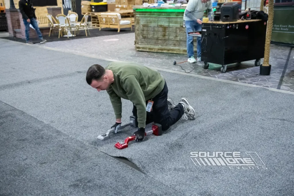 Event crew member kneeling on a convention center floor, using carpet installation tools to align and secure gray flooring during trade show setup. Additional staff, furniture, and event equipment are visible in the background.