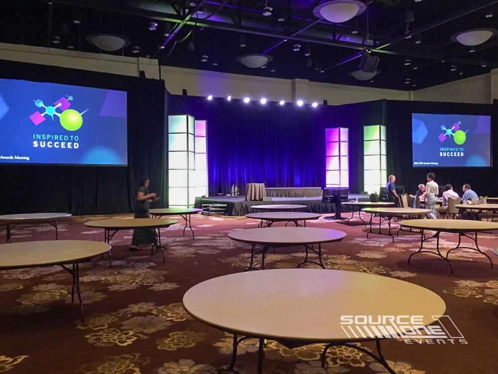 Elegant corporate presentation stage with purple accent lighting, circular seating, and branded conference graphics.