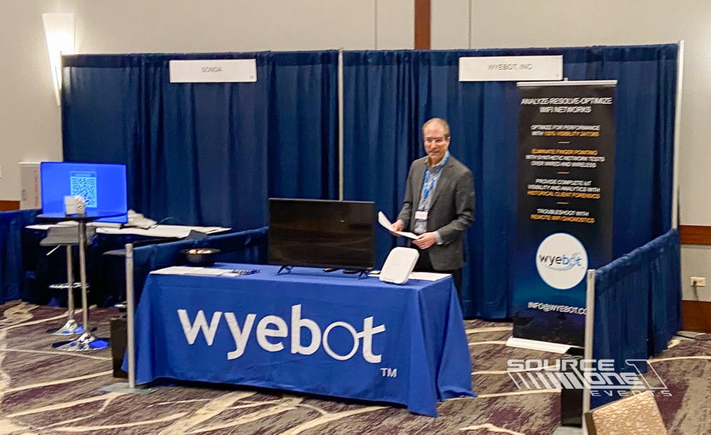 Wyebot branded exhibit table with custom table cover and presenter standing beside technology display for conference attendees.