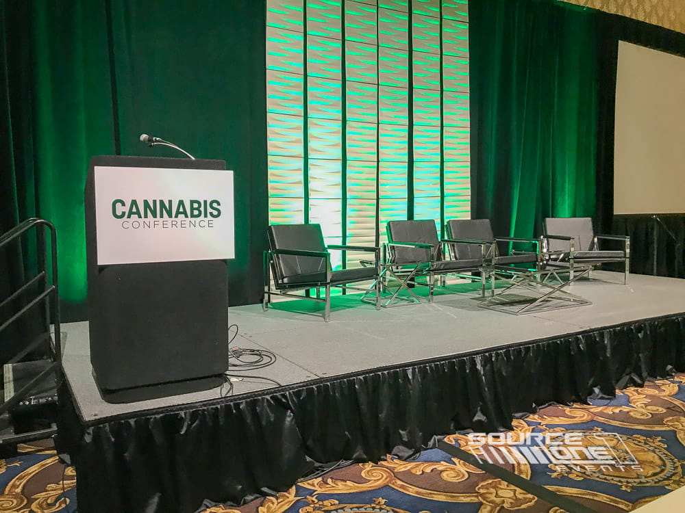 Modern conference stage featuring green illuminated backdrop, scenic elements, and presentation seating for industry education.