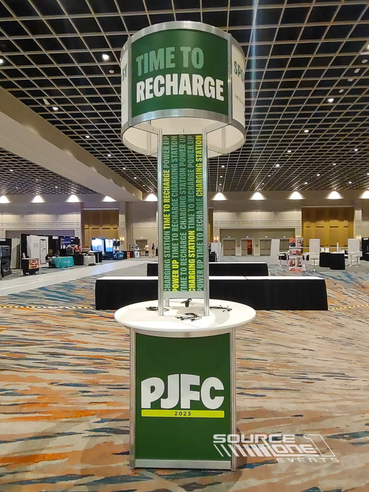 Cylindrical charging tower wrapped in vibrant green branding with multiple device charging ports positioned on a convention show floor.