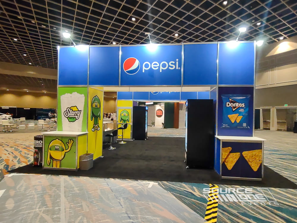 Custom 20x20 Pepsi trade show booth featuring bold blue branding, illuminated overhead signage, beverage displays, and interactive product sampling space designed by Source One Events.