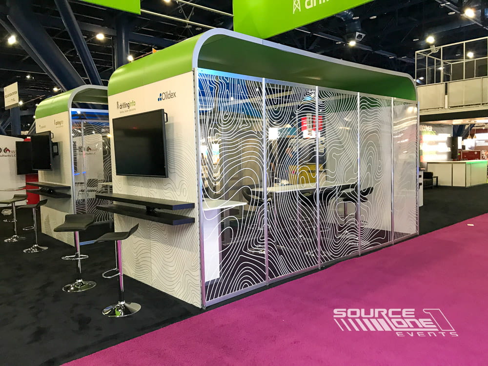 Large custom island exhibit featuring green overhead branding, glass meeting rooms, digital displays, and premium exhibit construction.