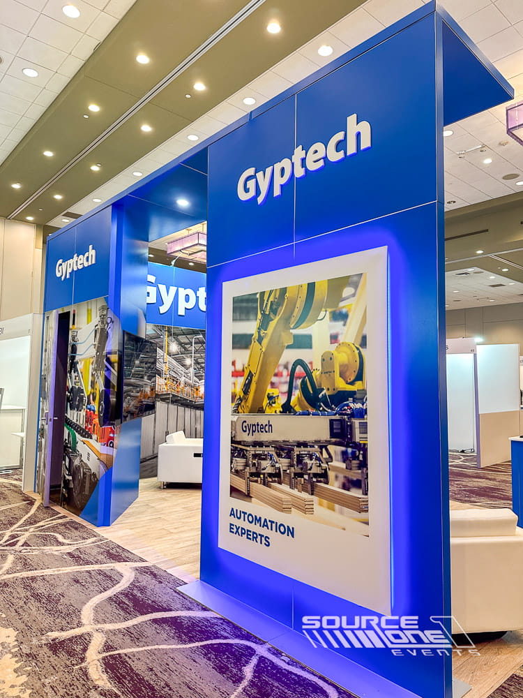 Large industrial trade show exhibit with blue illuminated branding, product displays, and collaborative meeting environment.