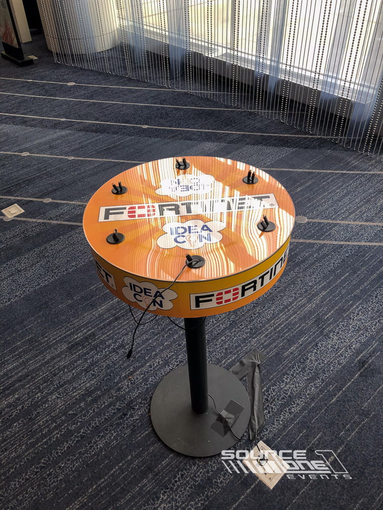 Circular charging table wrapped with colorful conference graphics, offering integrated power access and collaborative networking space.