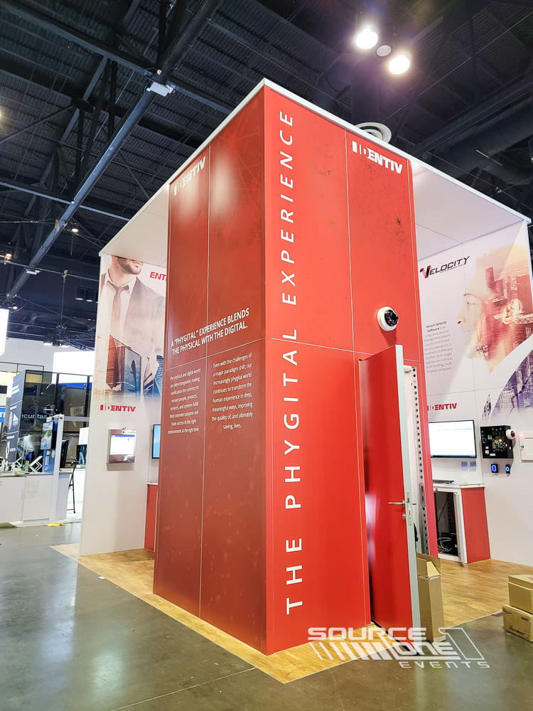 Large custom trade show booth featuring bold red architectural branding, overhead structures, and immersive product displays.