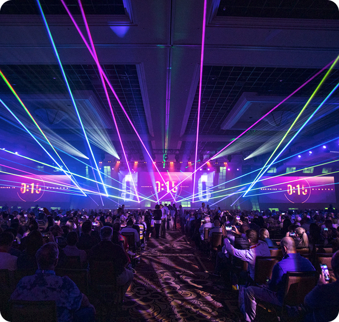 Concert-style general session with colorful laser lighting, stage production, and audience seating inside a large convention venue.