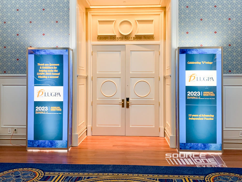 Pair of illuminated blue backlit towers framing ballroom entrances with elegant conference branding and directional messaging.