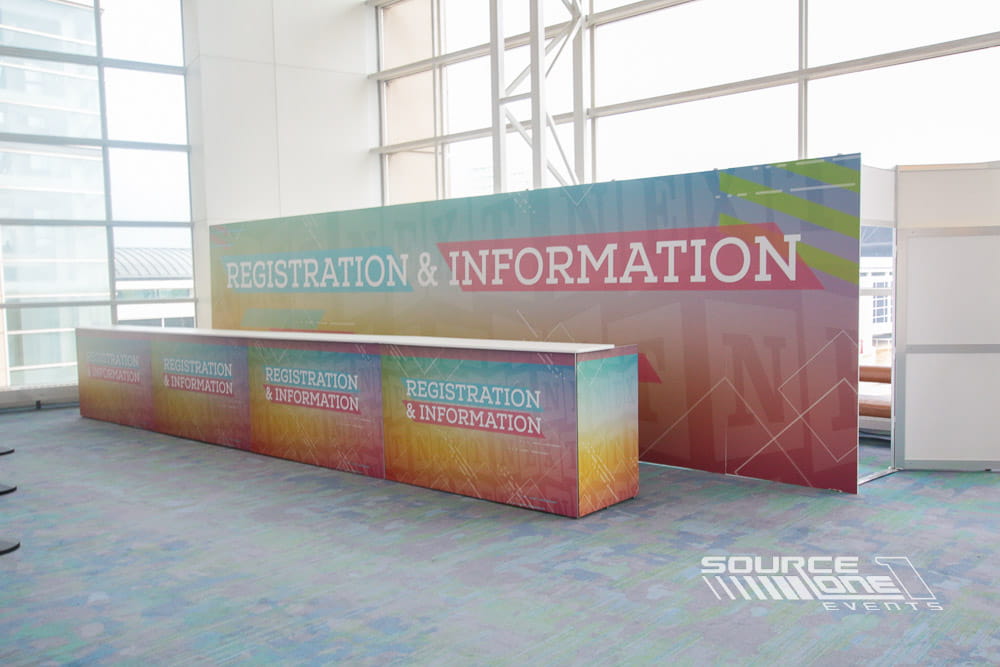 Modern registration counter featuring illuminated branded graphics, colorful information panels, and attendee check-in stations.
