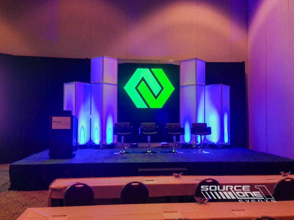 Contemporary keynote stage with blue uplighting, illuminated branded graphics, and theater seating for conference attendees.