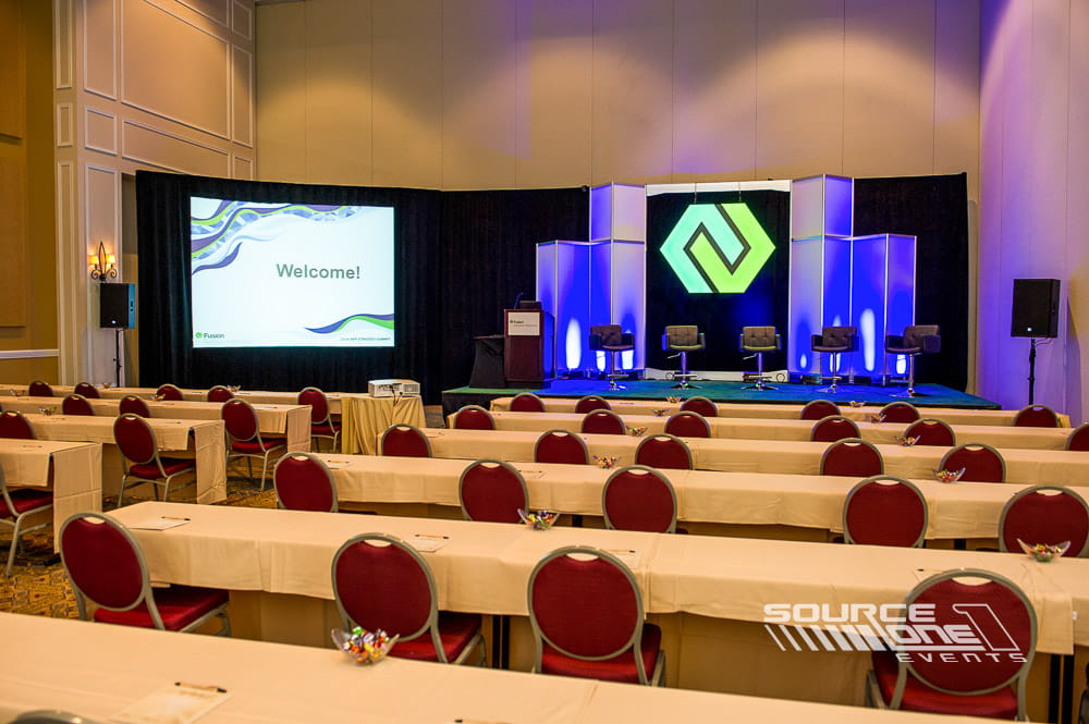 Large conference stage featuring multiple projection screens, blue lighting effects, and professional audiovisual production.