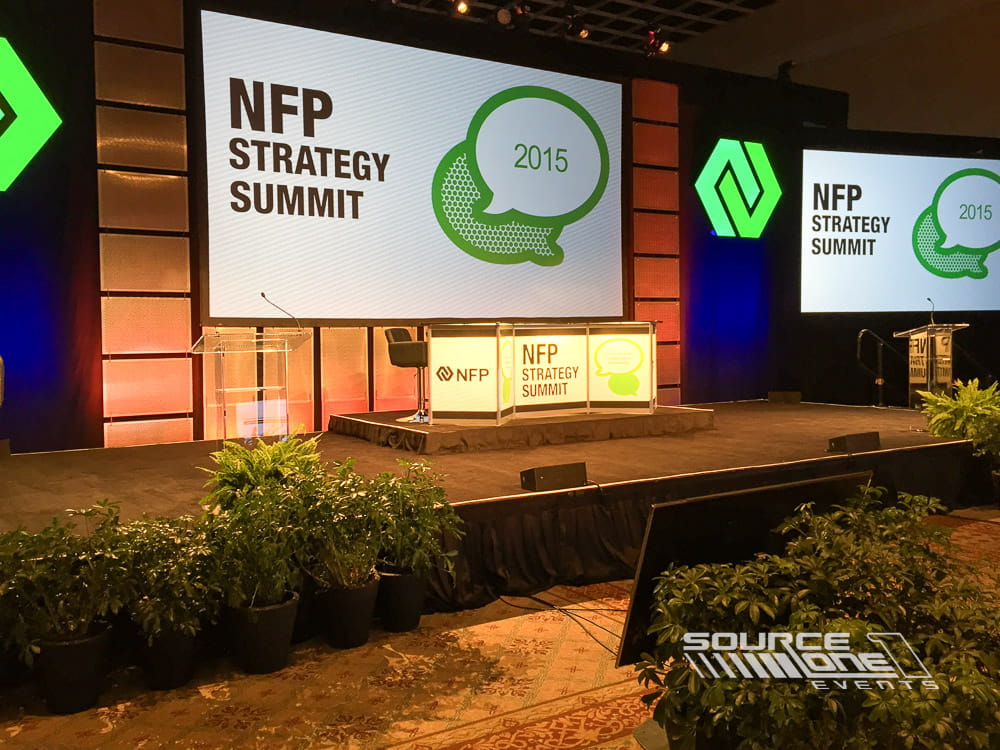 Corporate conference stage featuring green LED screens, branded presentation graphics, and modern stage production for keynote sessions.