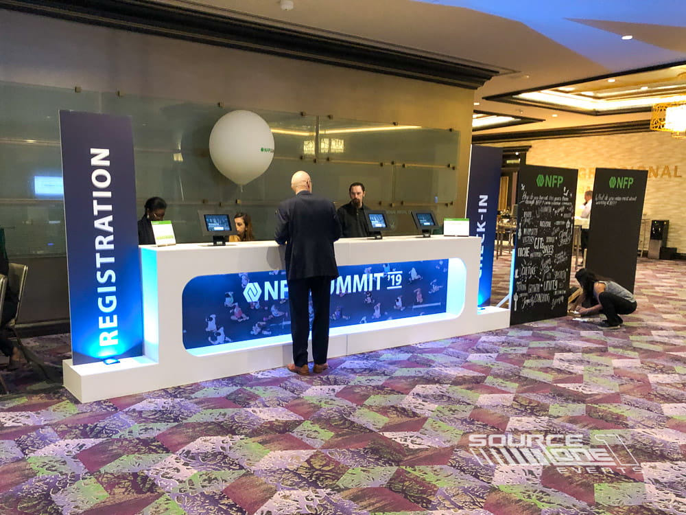 Contemporary registration desk with digital displays, blue branding, and multiple attendee check-in stations.