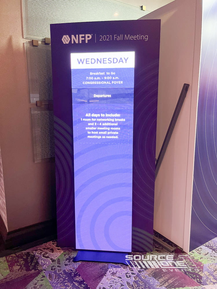 Tall digital LED information display featuring purple conference branding and attendee wayfinding graphics.