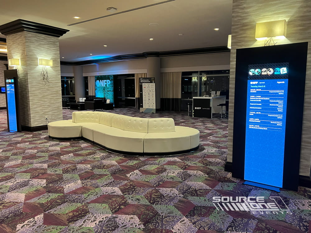 Contemporary networking lounge featuring illuminated LED poster display, white seating, and modern conference branding.