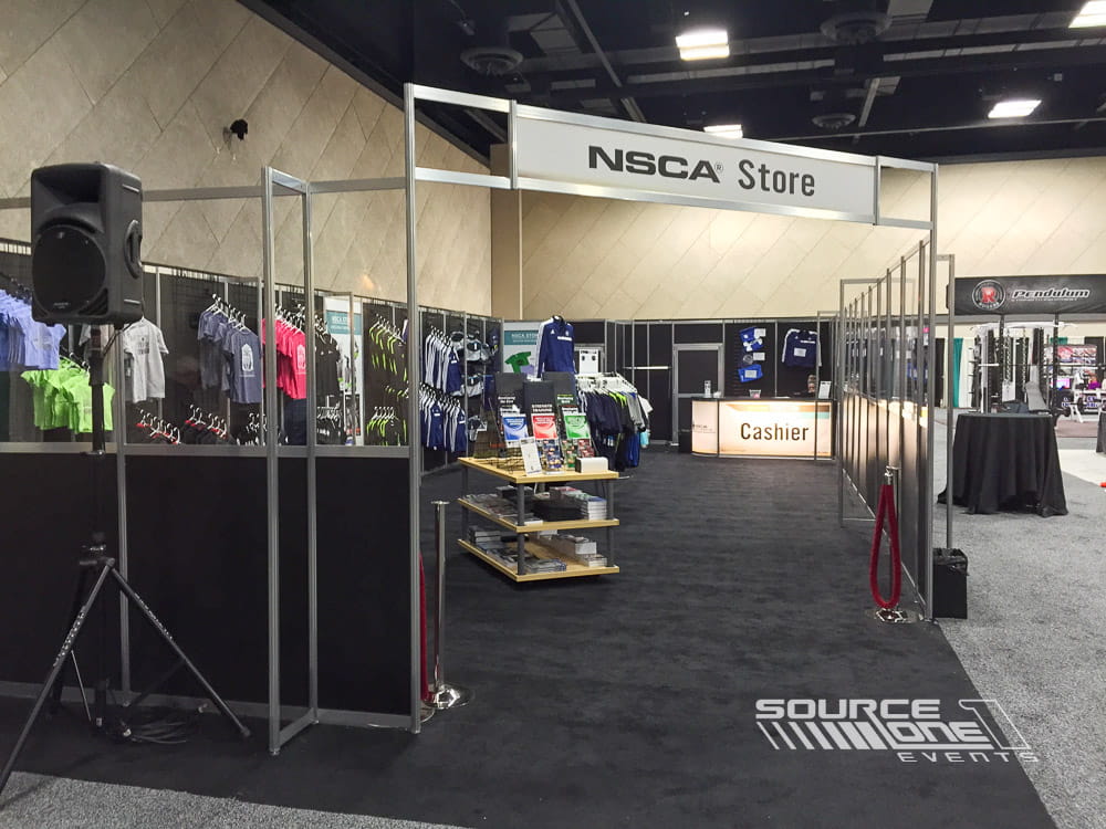 Wide entrance to conference retail store with overhead branding, product displays, and attendee shopping experience.