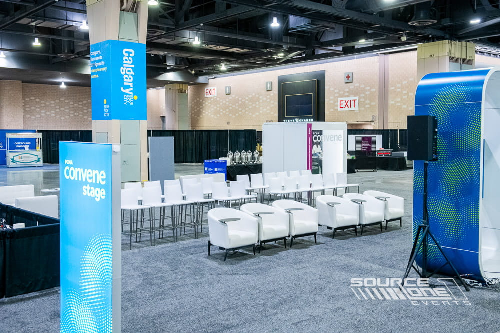 Contemporary conference presentation stage with white modular seating, branded educational graphics, and open networking environment.
