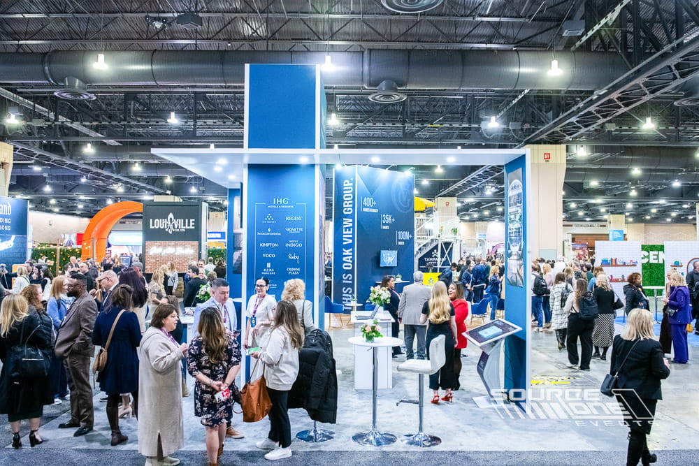 Large custom IHG exhibit featuring illuminated blue branding, overhead structures, meeting spaces, and premium exhibit fabrication on the trade show floor.