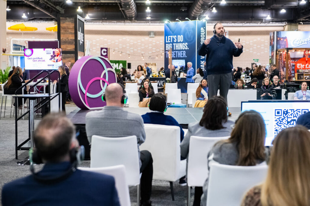 Speaker presenting to attendees seated in a conference theater on the trade show floor, surrounded by exhibitor booths and interactive event experiences.