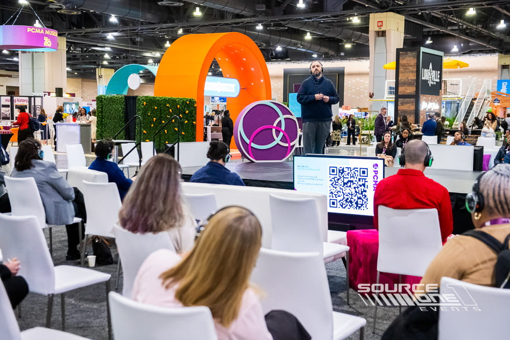 Interactive educational stage with circular architectural elements, branded presentation screens, audience seating, and collaborative learning environment.