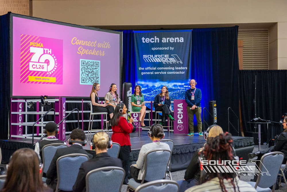 Contemporary educational stage featuring vibrant pink and blue graphics, digital presentation screens, attendee seating, and open learning environment within the exhibit hall.