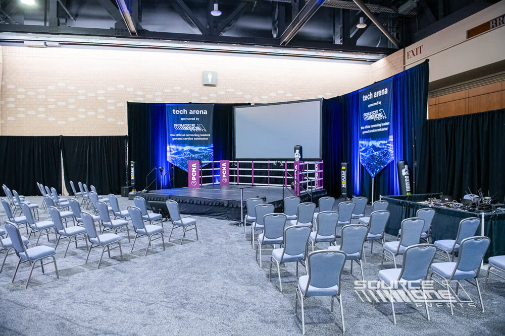 Modern technology presentation stage with blue branded graphics, integrated LED displays, white seating, and interactive learning space at PCMA Convening Leaders.
