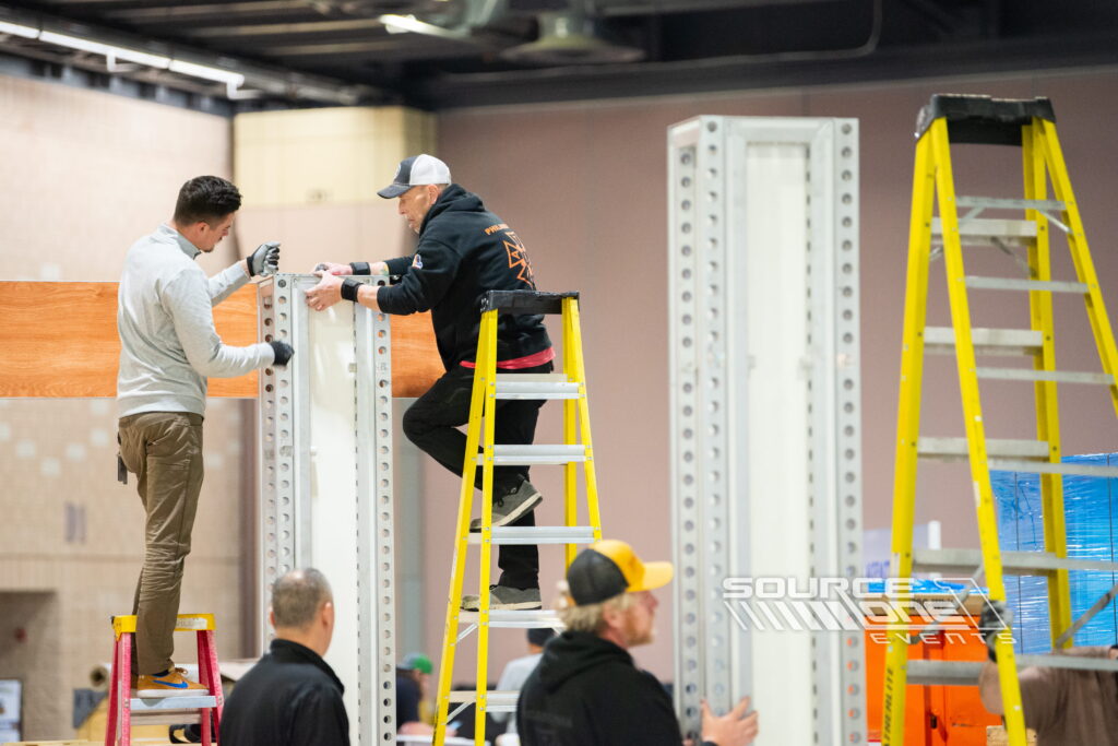Crew member installing exhibit components using a ladder during event setup.