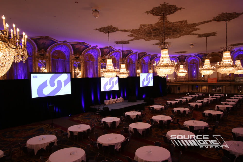 Elegant conference stage with purple and amber uplighting, branded presentation screens, decorative chandeliers, and theater seating for keynote sessions.