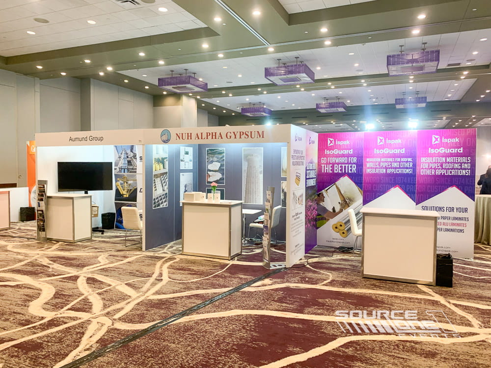 Large open exhibit booth with blue branding, white furniture, digital displays, and collaborative networking space.
