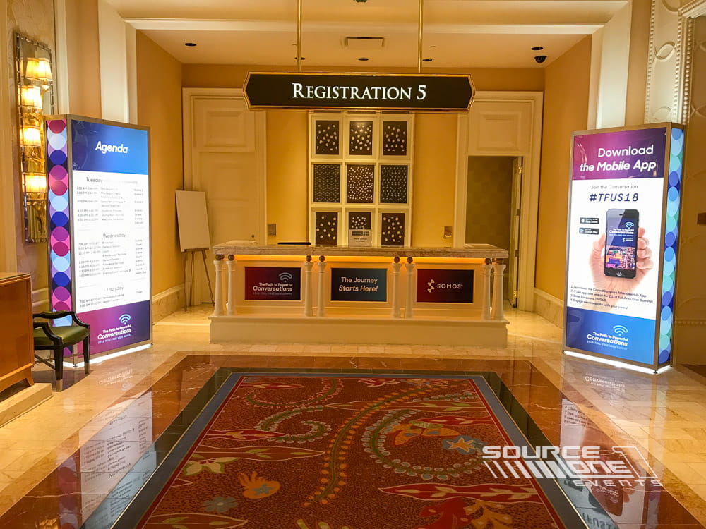 Registration entrance featuring illuminated backlit tower displays with warm wood accents and branded conference graphics.