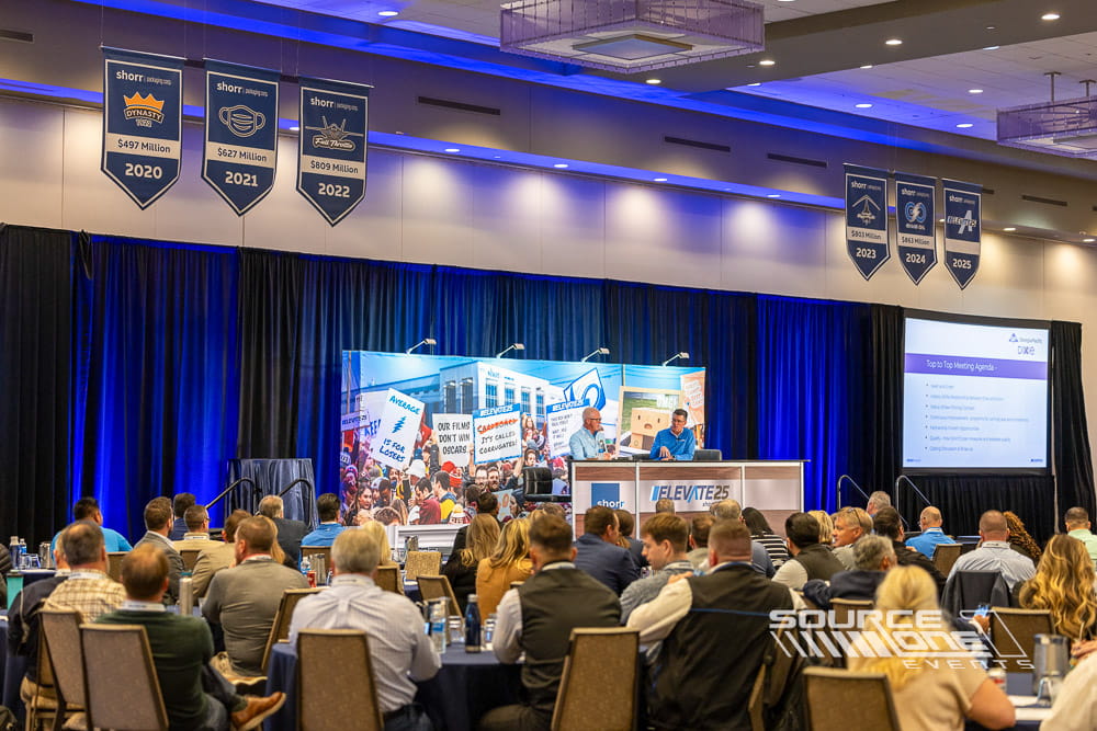 Large general session stage featuring blue LED lighting, dual presentation screens, branded graphics, and theater-style audience seating at a corporate conference produced by Source One Events.