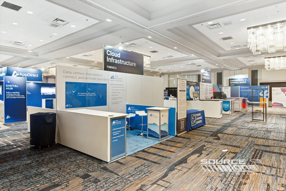 Large open exhibit booth with blue branding, white furniture, digital displays, and collaborative networking space.