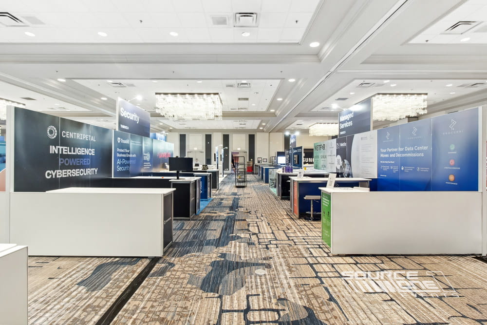 Bright trade show booth with overhead branding, multiple exhibit stations, integrated monitors, and open attendee circulation.