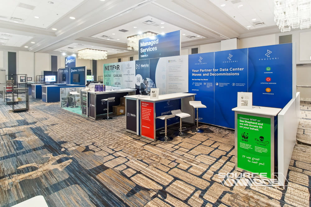 Spacious custom exhibit featuring blue branding, digital signage, meeting tables, and premium hardwall architecture.