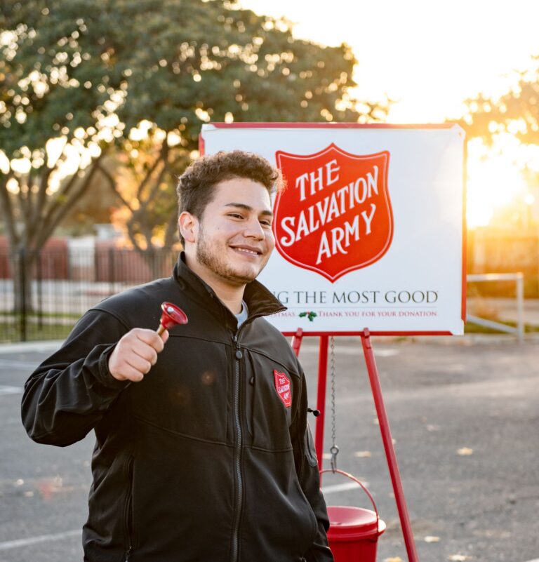 A cheerful volunteer ringing the Salvation Army bell outdoors during the day.