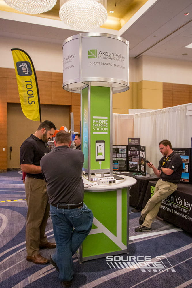 Green branded mobile charging station installed within a trade show floor to provide attendee device charging and sponsor visibility.